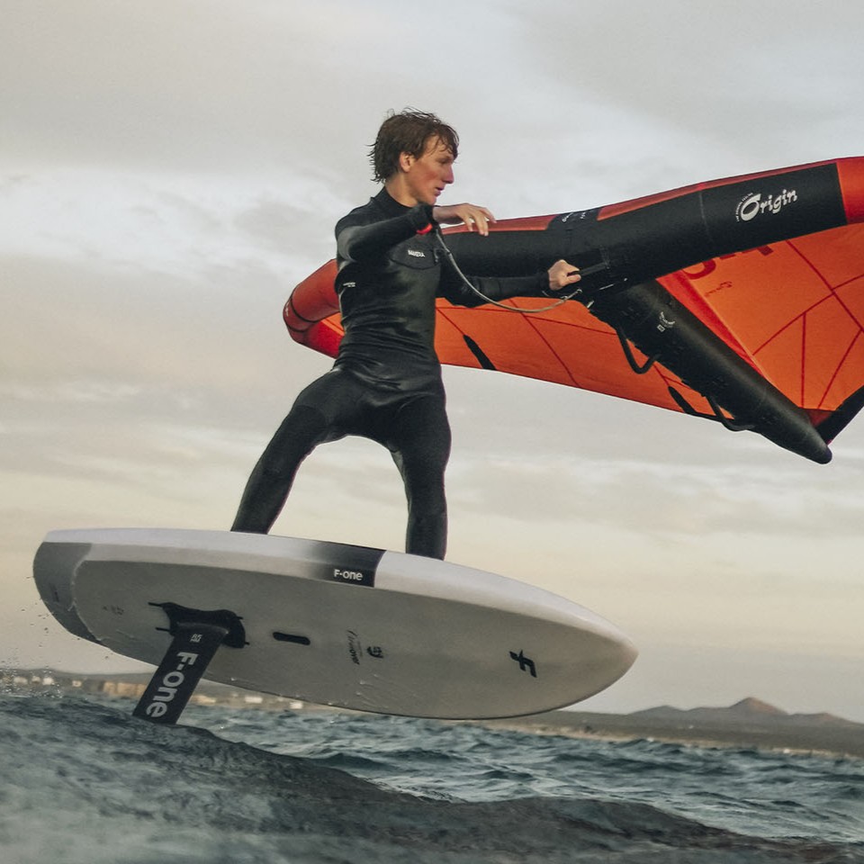 Rider en action sur la F-One Rocket Wing Crossover, planche de wingfoil polyvalente pour le surf, le wing et le dockstart, équipée d’un foil F-One, en vol au-dessus de l’eau avec une aile orange F-One Origin.
