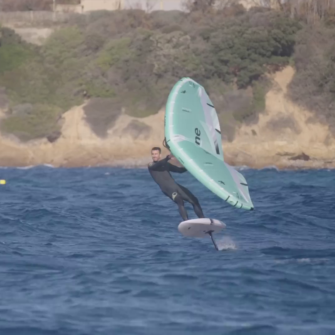 Rider en combinaison noire pratiquant le wingfoil avec une aile Strike V5 turquoise sur une mer agitée, devant une côte rocheuse et végétalisée.