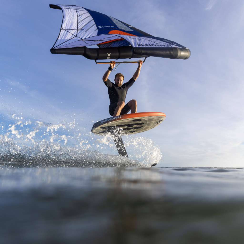 Rider en combinaison noire pratiquant le wingfoil sur une eau très calme, sous une météo ensoleillée.