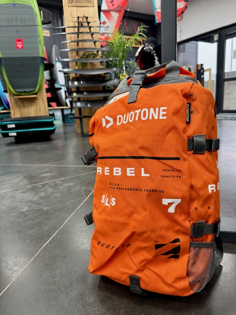 Sac de rangement orange pour la Rebel SLS 2025, avec une boutique de sports nautiques en arrière-plan.